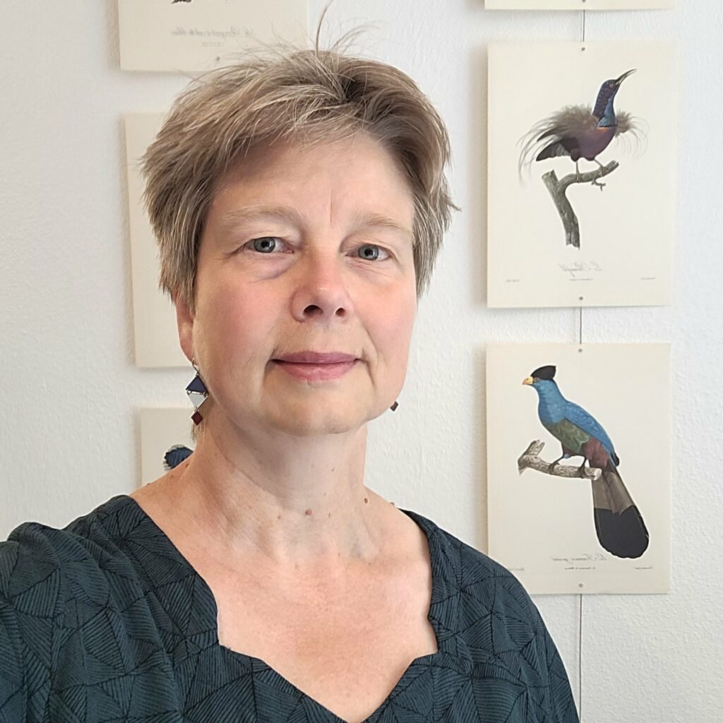 Porträtfoto einer mittelalten Frau mit kurzem graublondem Haar und einer dunkelblauen, gemusterten Bluse; dahinter an der Wand Stiche von Paradiesvögeln